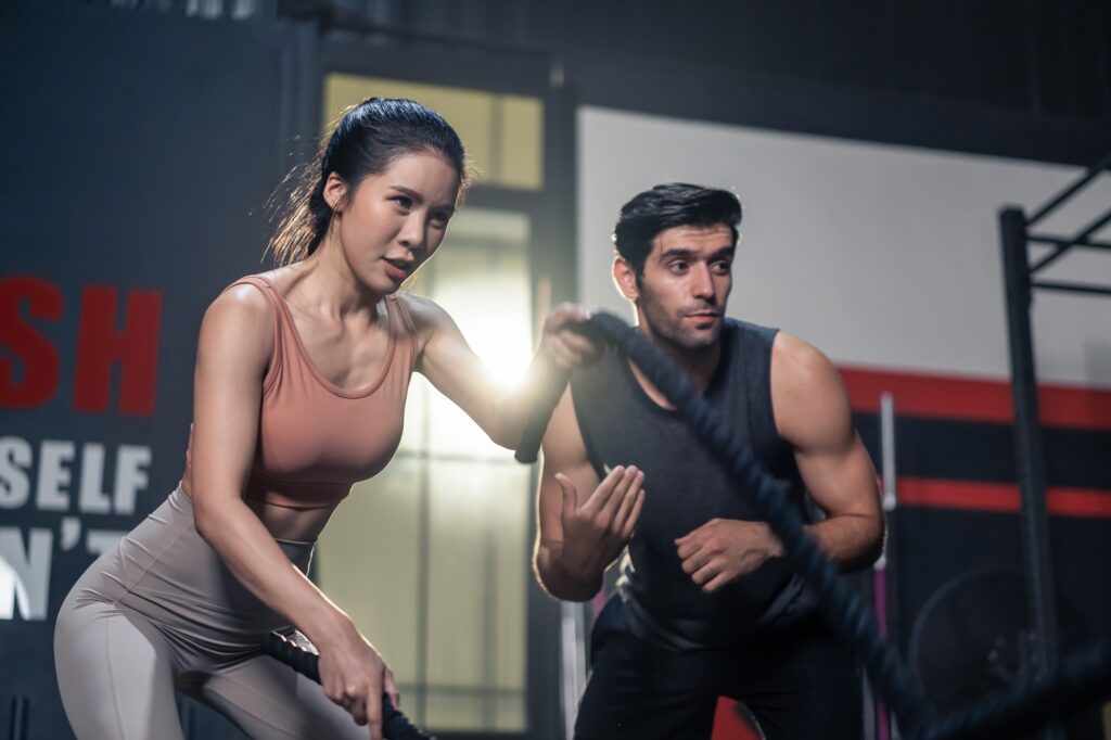 Caucasian Fitness trainer motivate young active beautiful sport girl using battle Rope in gym